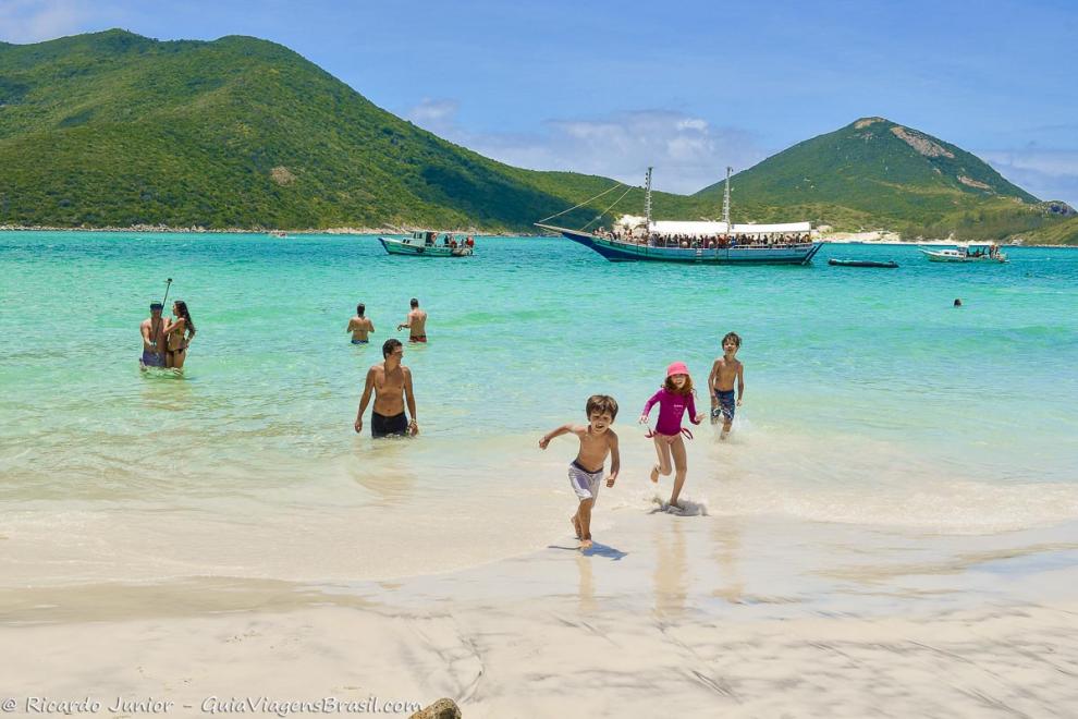 Imagem de crianças brincando na beira da praia e casal tirando fotos no mar em Arraial do Cabo.